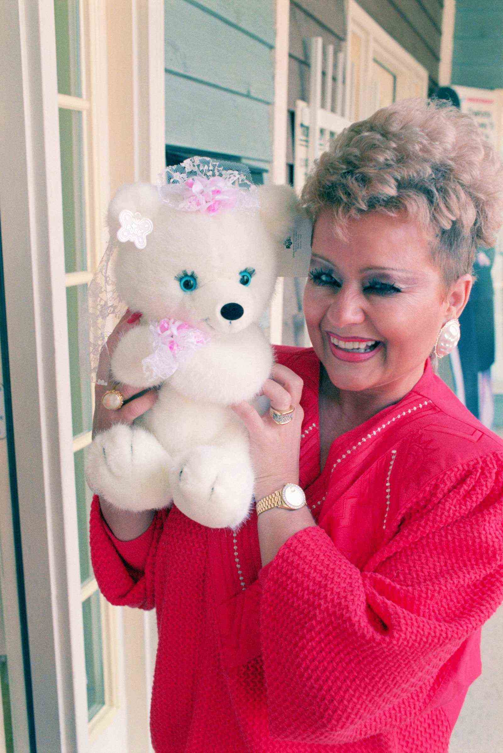 Tammy Faye Bakker trägt einen leuchtend roten Pullover und einen blauen Lidschatten und hält einen ausgestopften weißen Bären in die Kamera
