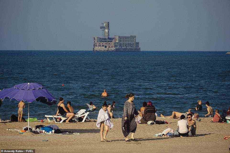 Eine Strandszene in Kaspiysk in Dagestan, die an der Küste des Kaspischen Meeres liegt.  Im Hintergrund ist ein verlassenes Marinewaffenwerk von 1931 zu sehen, das eine Torpedowerkstatt beherbergte