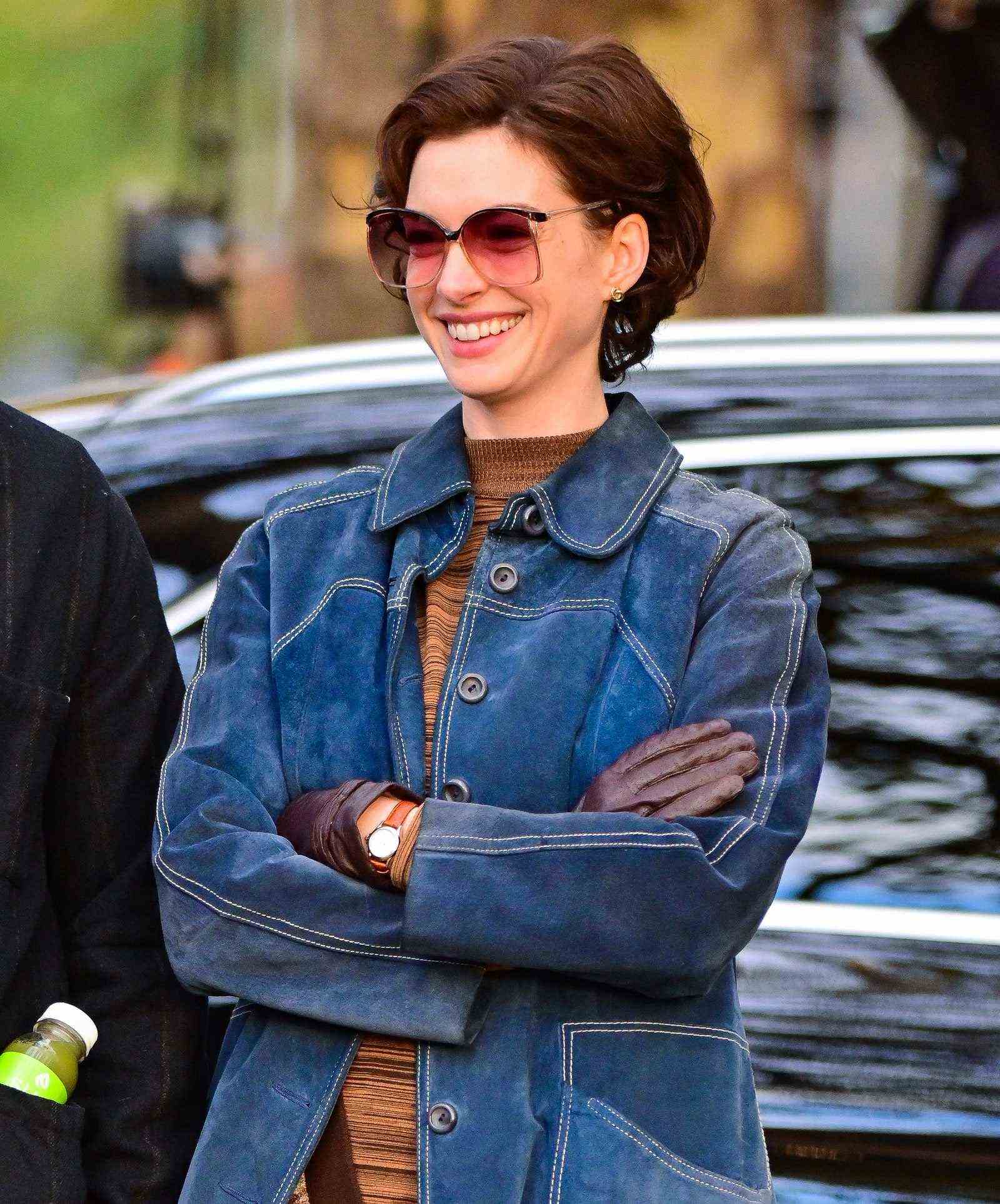 Anne Hathaway lacht am Set von Armageddon Time und trägt eine Jeansjacke und eine Sonnenbrille mit einem kurzen lockigen Bob