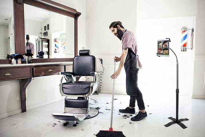 Picture shows a man in a barber shop sweeping hair off the floor while his tablet rests in an upright CTA Digital height-adjustable gooseneck stand.