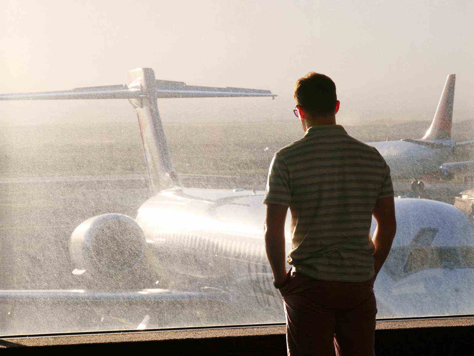 Jake Lacy steht an einem Flughafenfenster und schaut auf die Flugzeuge.