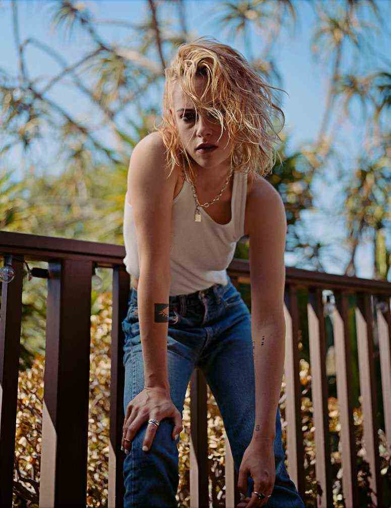 Kristen Stewart in Jeans und einem weißen Tanktop mit unordentlichen gebleichten Haaren beugt sich draußen vor und schaut direkt in die Kamera.