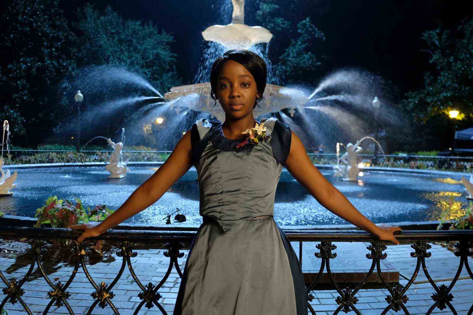 Thusu Mbedu trägt ein blaues Partykleid und lehnt sich an ein Geländer vor einem großen Brunnen.