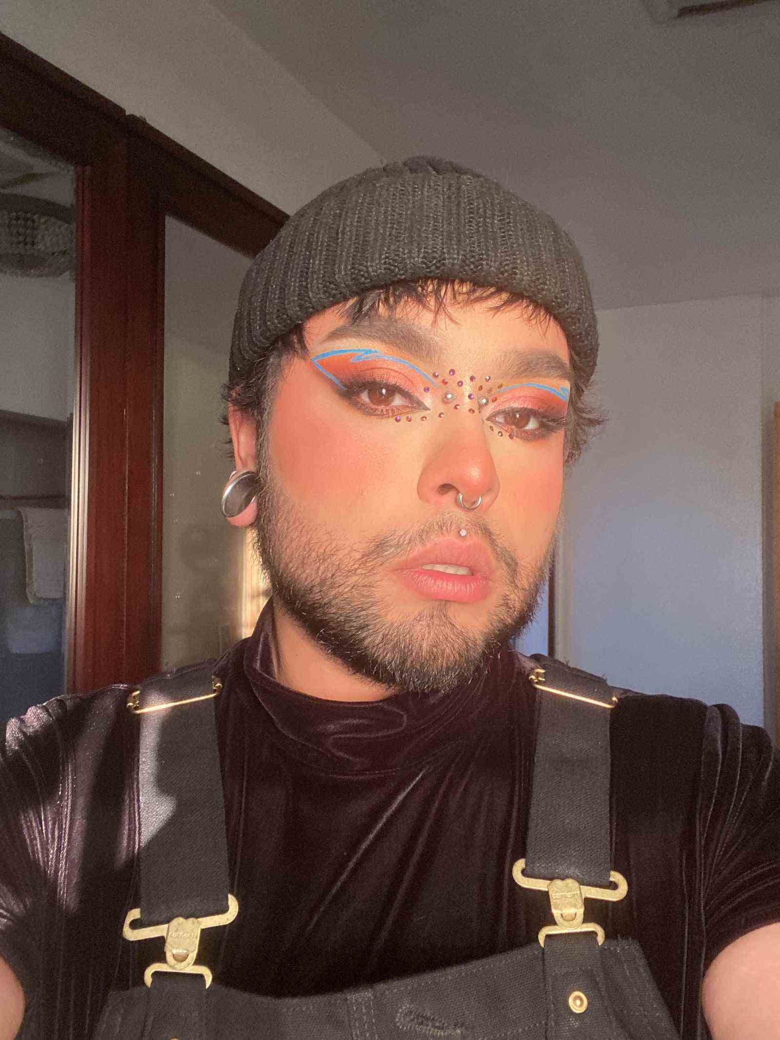 Selfie des nicht-binären Beauty-Influencers Realsickpuppy mit dramatischem Augen-Make-up in Blau und Pink, einer grauen Mütze und schwarzem ...