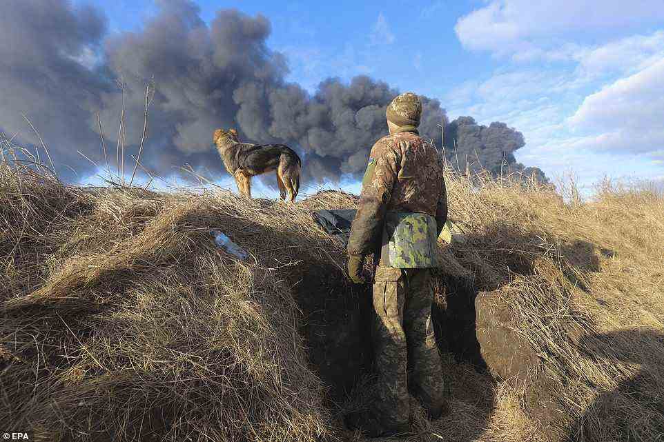 A Ukrainian serviceman and his dog stands in a position looking at smoke from a burned petroleum storage depot behind after a Russian missile attacked near Kyiv