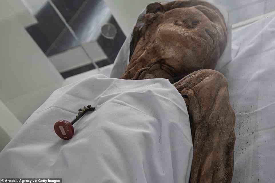 The mummy also has a cross resting on top of the chest and white linen over the head, which is turned to the side of the glass case