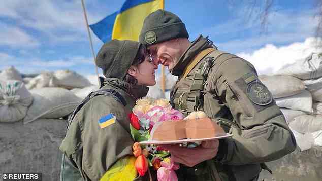 Dem Paar werden ein traditioneller Kuchen und Blumen überreicht, während im Hintergrund eine ukrainische Flagge weht