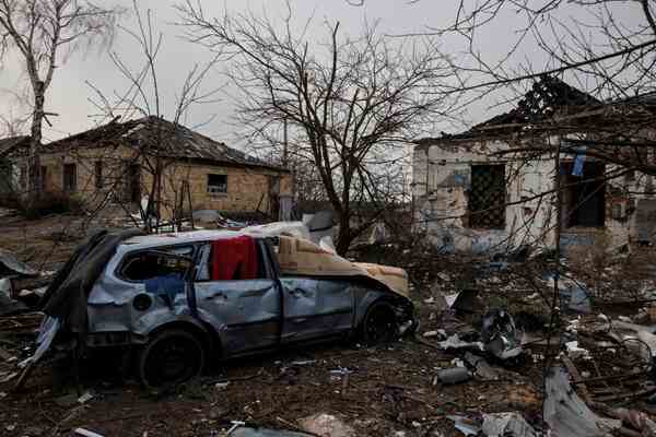 A destroyed car in front of two houses full of holes.