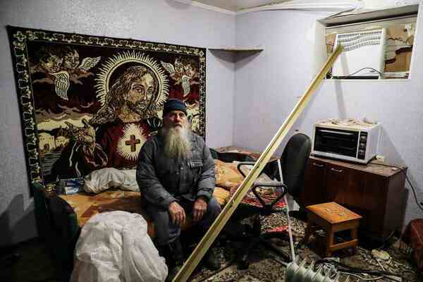 A man sits in a room with a Jesus tapestry behind and debris in front.
