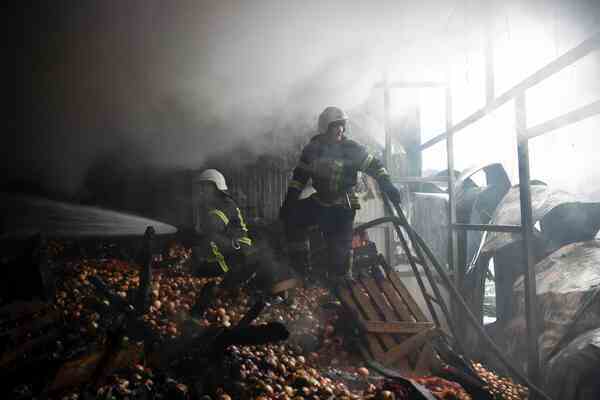Two emergency workers stand on a giant pile of onions. The air is smoky and part of the wall is open.