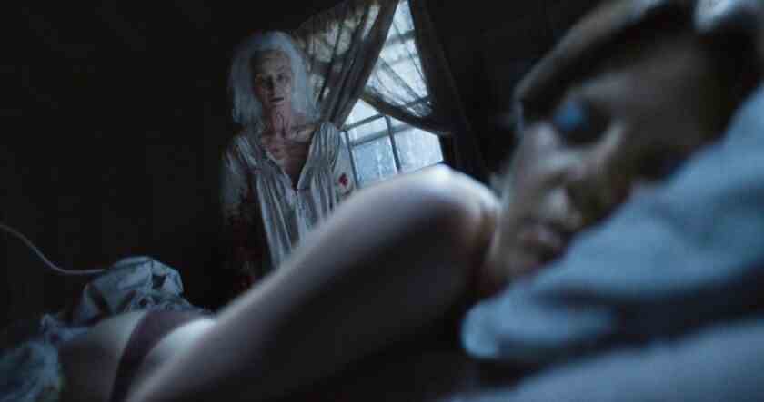 Im Horrorfilm liegt eine Frau auf einem Bett "X," mit einer alten Frau, die sie beobachtet.