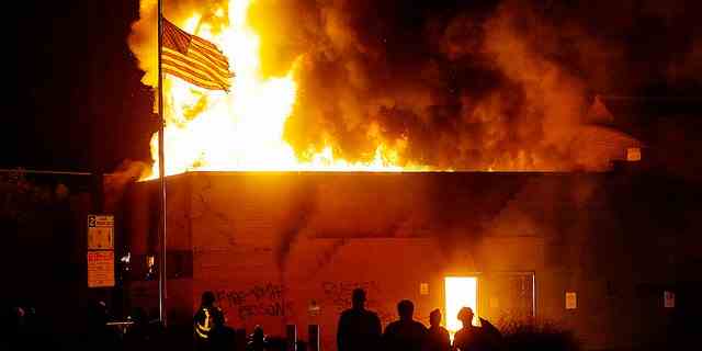 Menschen beobachten, wie die amerikanische Flagge während eines Aufstands, der gegen die Erschießung von Jacob Blake durch die Polizei am 24. August 2020 in Kenosha, Wisconsin, protestiert, über einem brennenden Gebäude weht. 