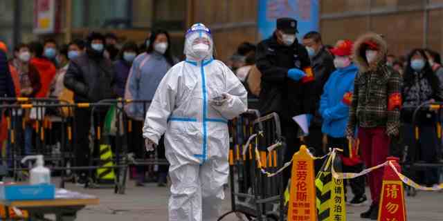 Ein Gesundheitspersonal, das einen Schutzanzug trägt, geht an maskierten Bewohnern vorbei, die an einem Coronavirus-Teststandort in einer Schlange warten, um ihren Rachenabstrich zu erhalten, nachdem am Mittwoch, dem 6. April 2022, in Peking ein COVID-19-Fall in einem Wohngebäude entdeckt wurde.