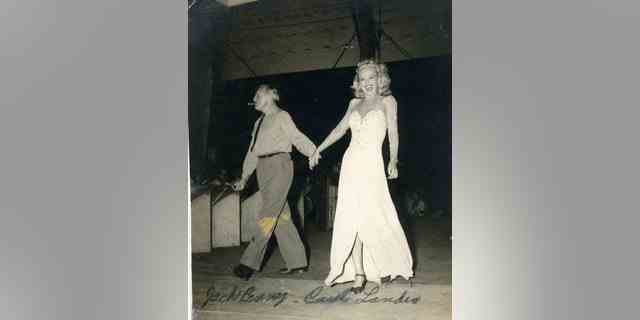 Carole Landis in einem weißen bodenlangen Kleid hält die Hand von Jack Benny.