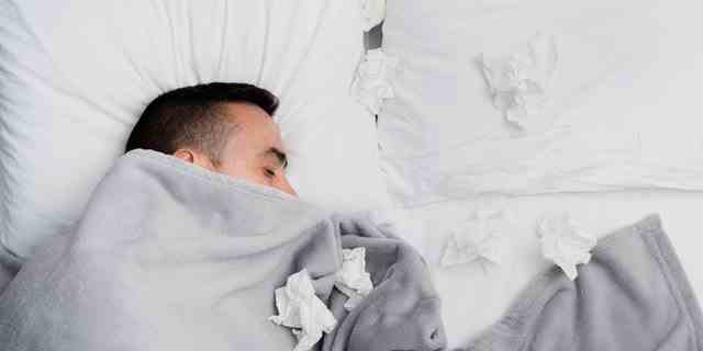 Kranker junger Mann im Bett, bedeckt mit einer hellgrauen Decke, umgeben von gebrauchten Geweben