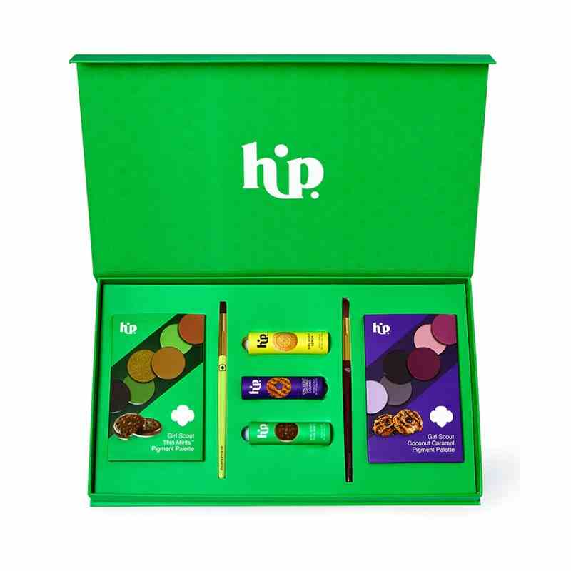 Ein weißer Hintergrund mit einer grünen Box darüber, die mit allen Make-up-Produkten aus der Kollaborationskollektion von HipDot X Girl Scouts gefüllt ist, darunter zwei Lidschatten-Paletten, zwei Make-up-Pinsel und drei Lippenstifte. 