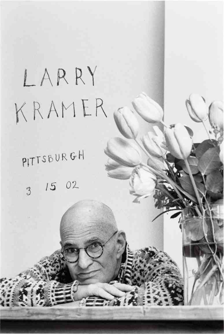 Larry Kramer stützt sein Kinn neben einem Strauß Tulpen auf die verschränkten Arme.