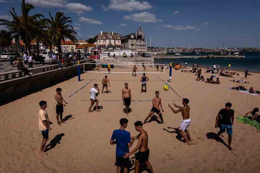 Menschen in Badehosen schlagen einen Volleyball in der Nähe eines Netzes.  Andere sonnen sich.  Im Hintergrund sind Gebäude und Palmen.