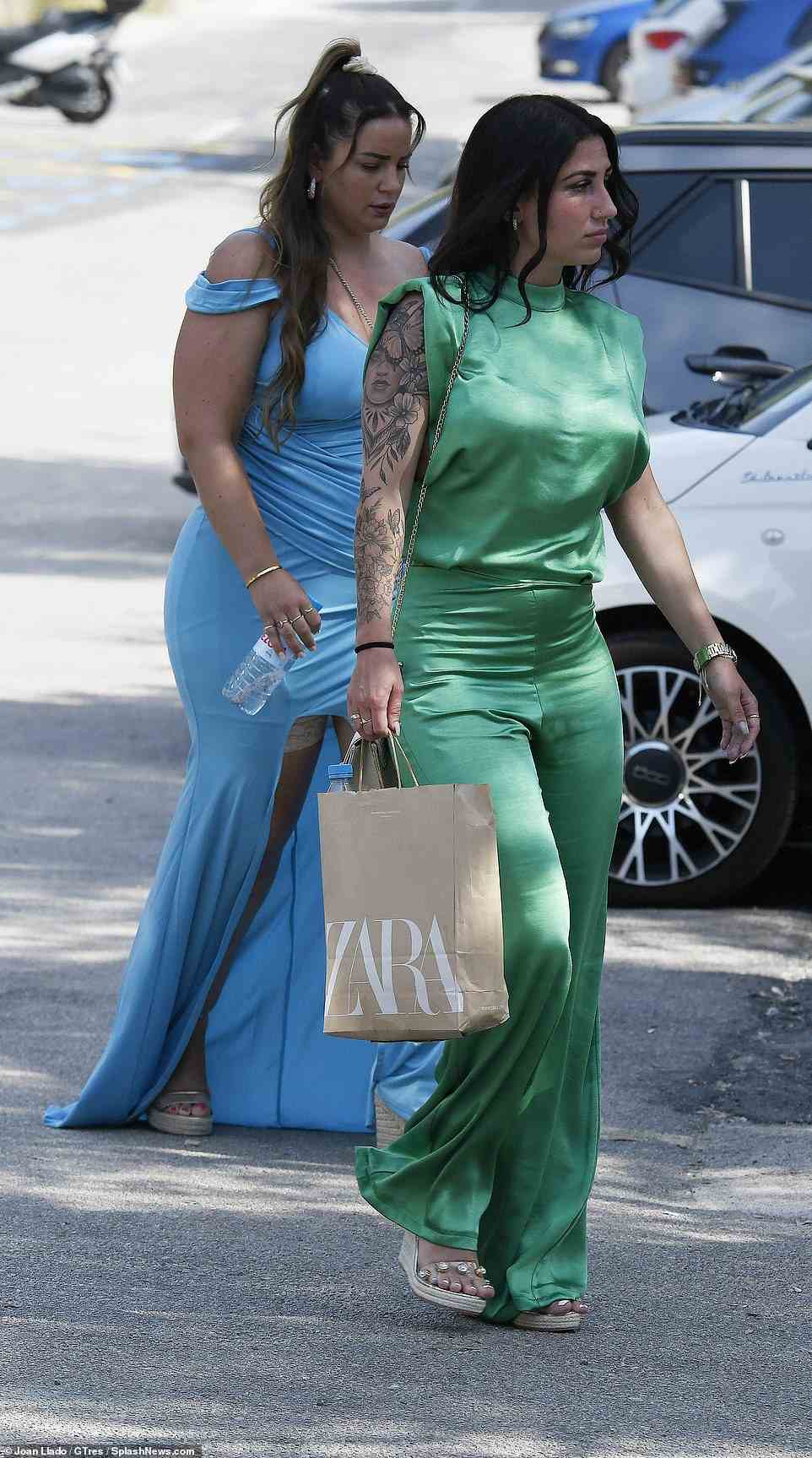 Ein Hochzeitsgast in einem eleganten, ärmellosen Katzenanzug aus grünem Satin und mit Keilabsatz, der eine Zara-Tasche trägt, betritt die Kulisse