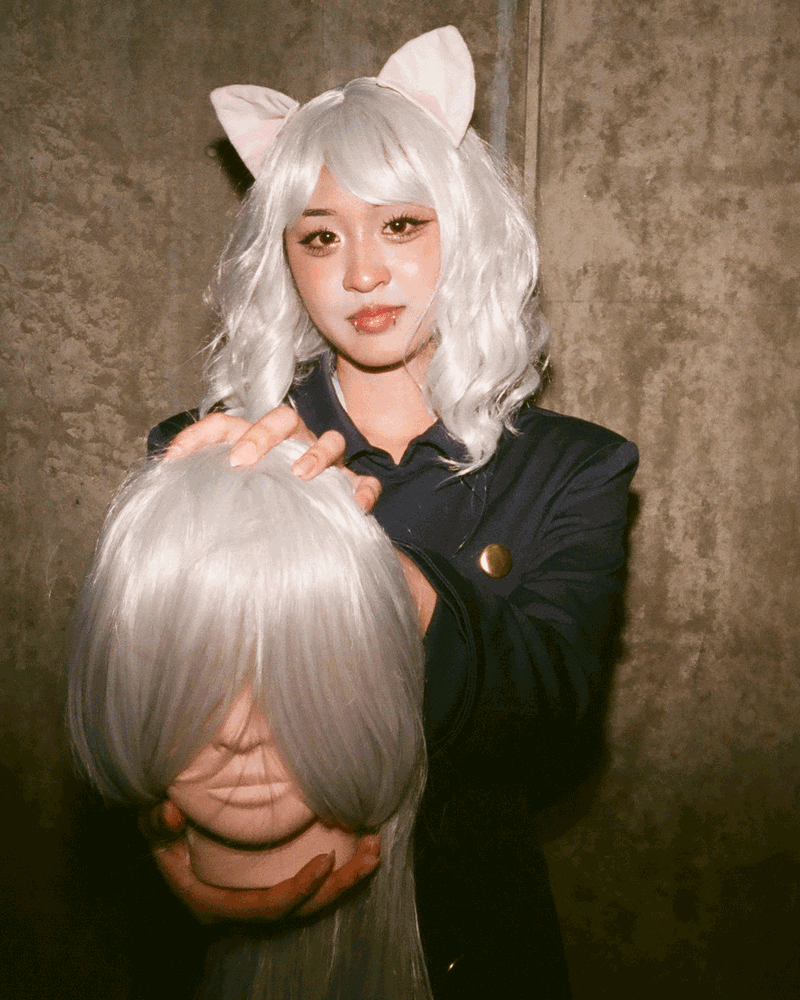 Eine Person, die eine blonde Perücke und Katzenohren trägt, hält einen gefälschten geköpften Kopf, ebenfalls mit blonden Haaren.