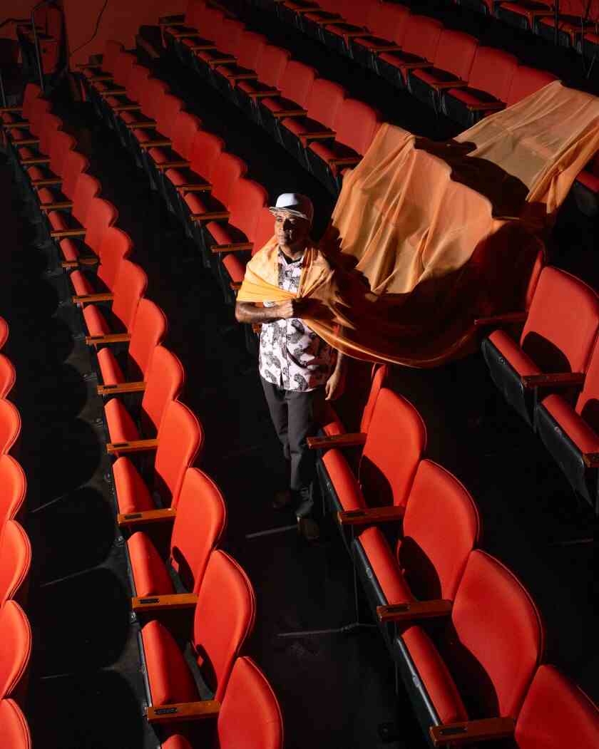 D'Lo in einem leeren Theater mit einem seidenen Stoffstreifen, der über zwei Sitzreihen hinter ihm schwebt.