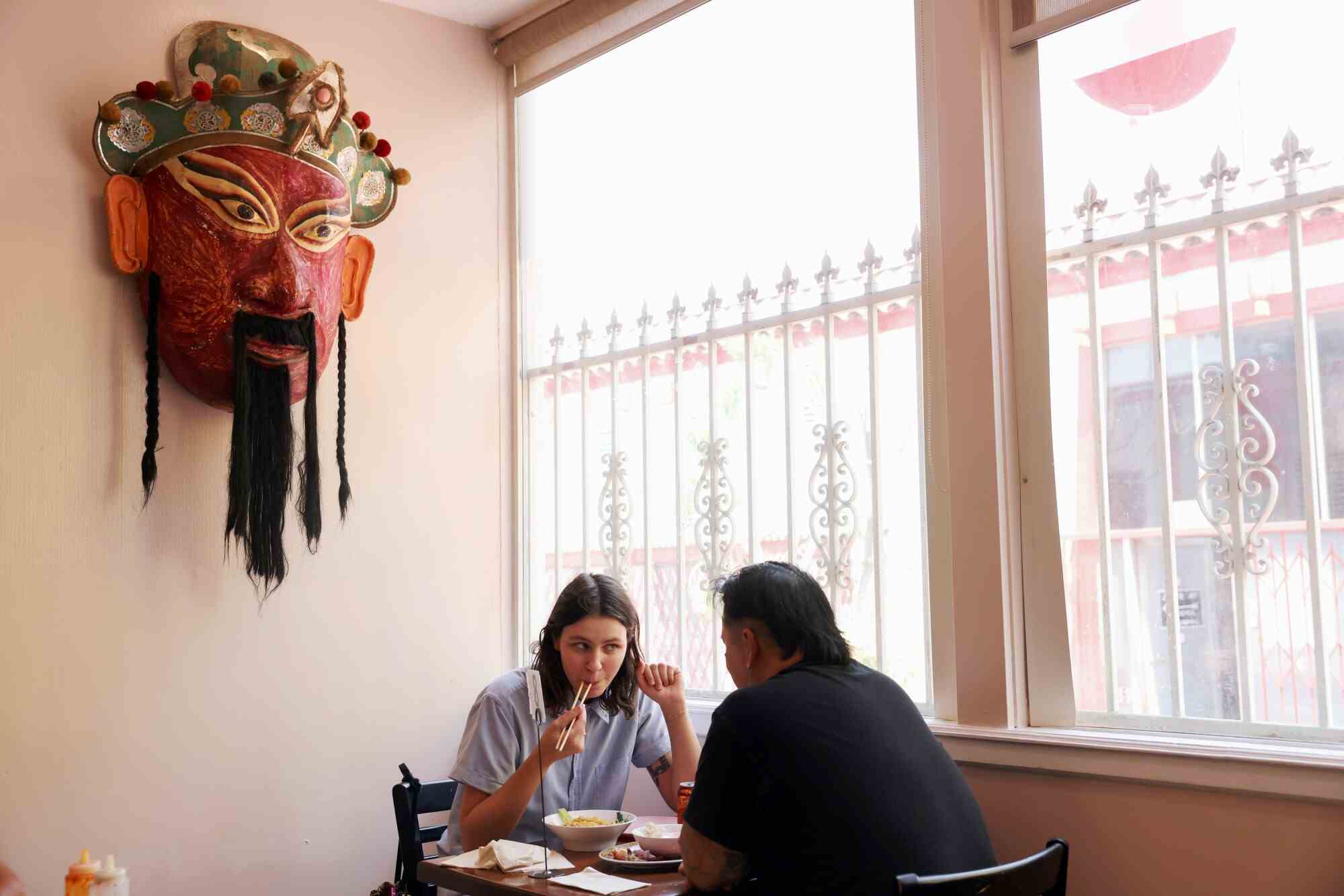 Zwei Personen speisen in einem Restaurant neben einem Fenster, über ihnen hängt eine große Maske an der Wand.