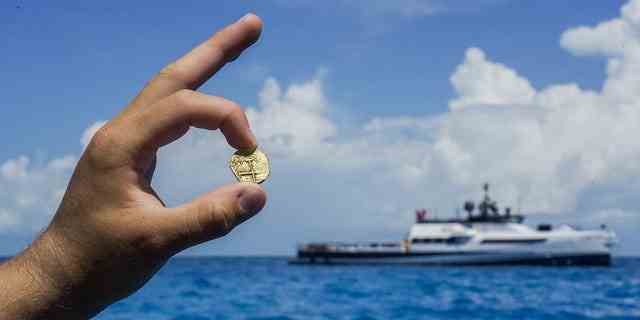 Ein Entdecker hält eine Goldmünze, die auf den Bahamas gefunden wurde, während ein Allen-Explorationsboot in der Ferne zu sehen ist.