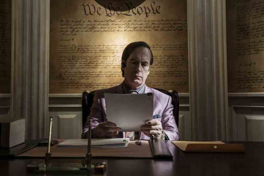 Ein Anwalt in einem rosafarbenen Anzug sitzt an einem hölzernen Schreibtisch und hält Papierkram in der Hand.  Ein Bild der Verfassung füllt die Wand hinter ihm.