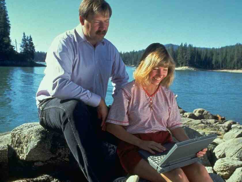 Ein Mann und eine Frau sitzen auf einigen Felsen und schauen auf einen alten Laptop, dahinter ein See und Bäume.