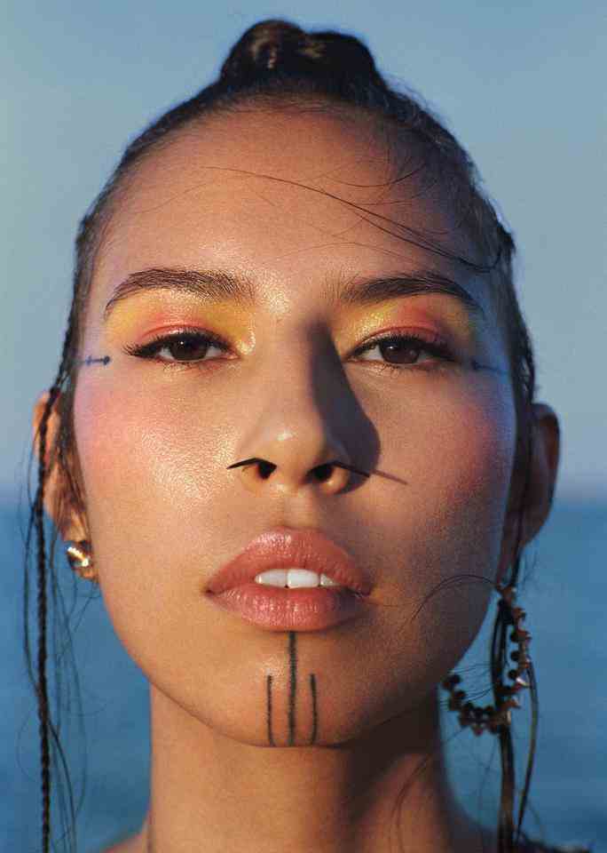 Close-up-Porträt von Model Quannah Chasinghorse, das direkt in die Kamera schaut.  Sie trägt sonnenuntergangsfarbenes Augen-Make-up, korallenrotes Rouge und passenden korallenroten Lippenstift.  Ihr Haar ist zu einem dünnen Dutt zurückgebunden. 
