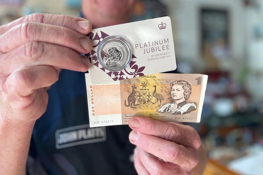 Nahaufnahme der Hände eines Mannes, die eine Gedenkmünze und eine 1-Dollar-Note mit dem Bild der Königin halten.