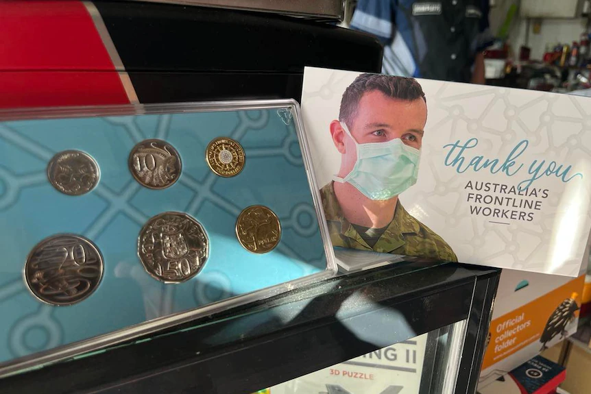 Ein Satz australischer Münzen in blauer Umgebung, neben einem Koffer mit einer Person mit Gesichtsmaske.