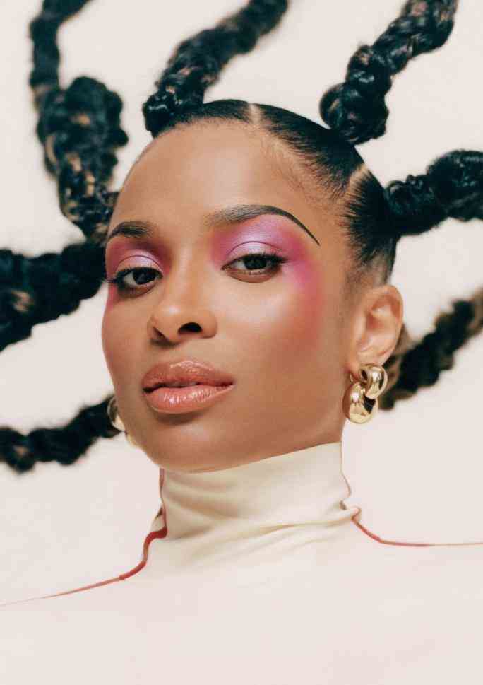 Close up portrait of Ciara wearing metallic pink eye makeup that drapes down into her cheekbones. Her hair is up in multiple braids that appear to be flying around her head.