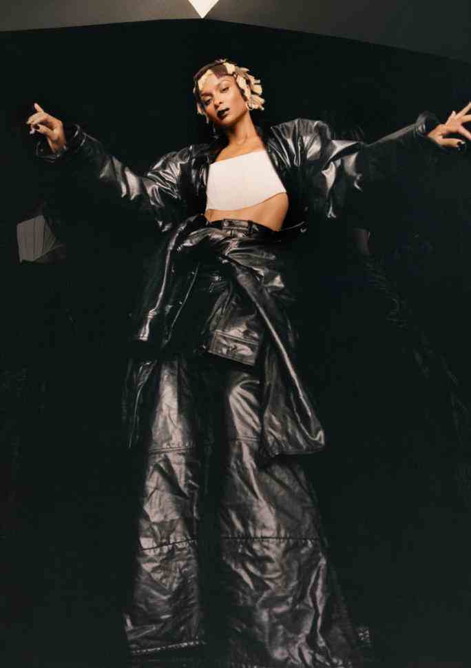 Full length portrait of Ciara wearing an oversized black jacket and black flare pants with a white bandeau top. She’s wearing a chin-length blonde wig with chunky brown lowlights. 