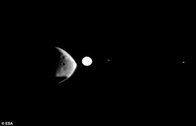 Der Marsmond Deimos (links), der sich durch seine felsige und mit Kratern übersäte Oberfläche auszeichnet, ist hier zu sehen, als er gerade am Jupiter vorbeizieht, der als großer weißer Fleck erscheint.  Jupiters Monde erscheinen als bloße Lichtflecken