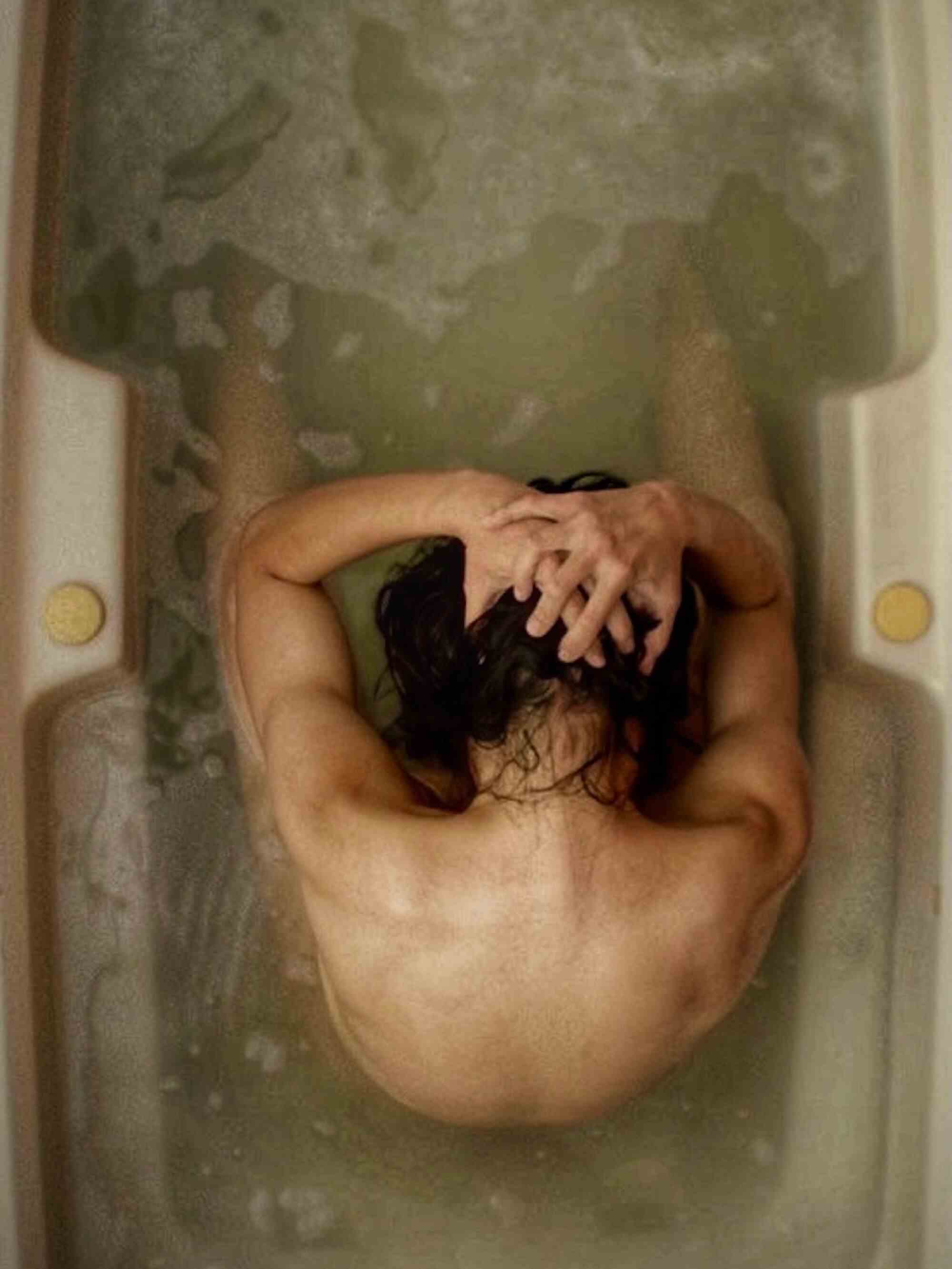 Eine Person von oben gesehen, sitzt nackt in einer Badewanne, die Hände auf dem Kopf.
