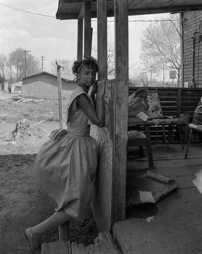 Eine junge Frau steht vor einem Holzhaus mit ihren Händen auf dem Rahmen der Veranda und blickt in die Kamera