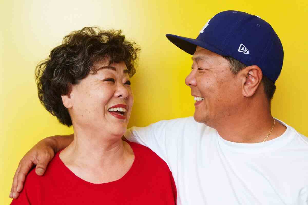 Jung Ye und Jeff Jun lachen und lächeln sich vor einer gelben Tür an.  Jeff hat seinen Arm um seine Mutter.