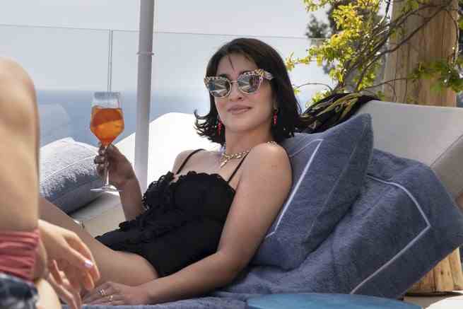 Simona Tabasco trägt als Lucia eine Sonnenbrille und lehnt sich mit einem Drink in der Hand auf einem Liegestuhl zurück.