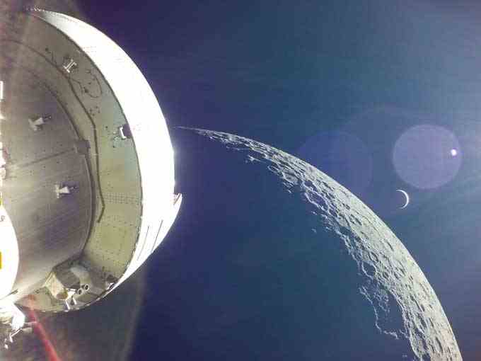 das Orion-Raumschiff im Vordergrund mit dem Mond im Hintergrund.  Die Erde wird als dünner Halbmond sowie als großer Linseneffekt mit einem winzigen Punkt in der Mitte dargestellt, der die Sonne darstellt