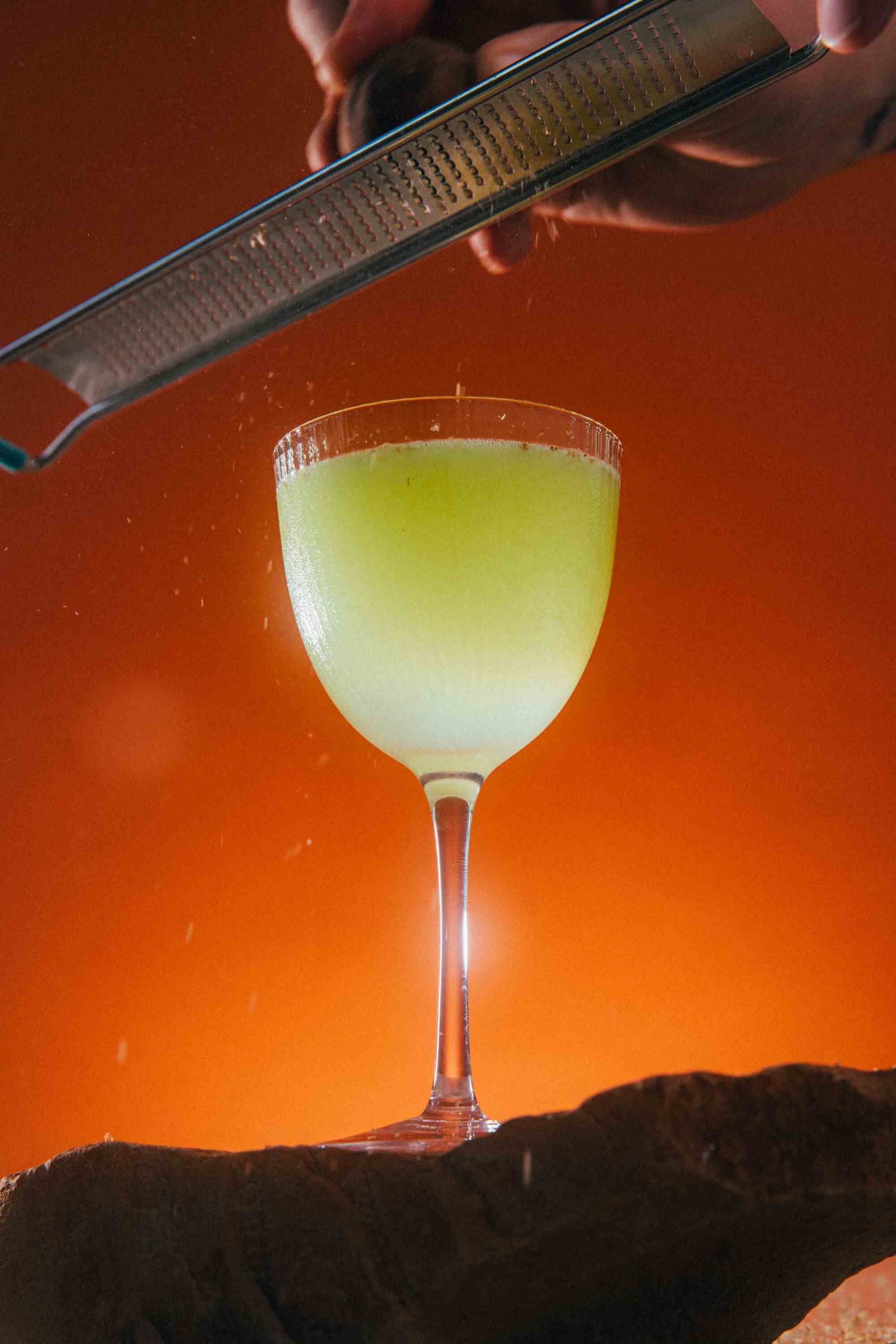 Ein grüner Cocktail leuchtet vor einem orangefarbenen Hintergrund.  Darüber reibt eine Hand die Schale einer schwarzen Limette in das Getränk.