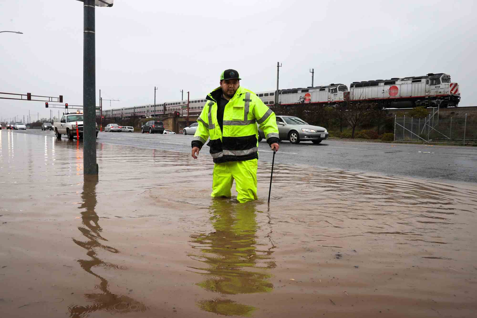 Ein Mann mit neongrüner Jacke und Hose steht knietief im schlammigen Hochwasser, während hinter ihm ein Auto und ein Zug vorbeifahren.