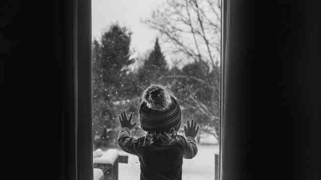 Schwarzweißfoto eines Kindes, das bei Schneefall aus dem Fenster schaut