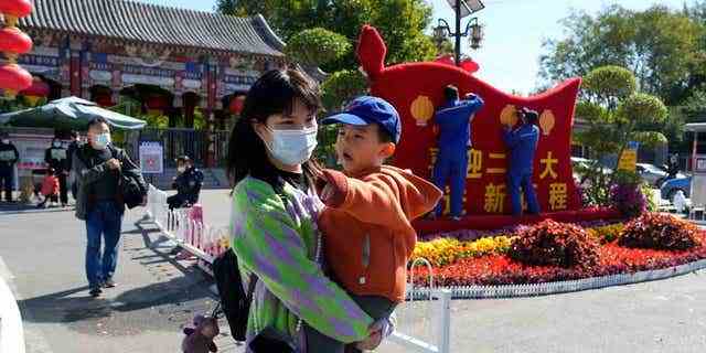 Eine Frau mit Maske trägt ein Kind an Arbeitern vorbei, die eine Ausstellung zur Feier des bevorstehenden 20. Parteitags in Peking dekorieren.