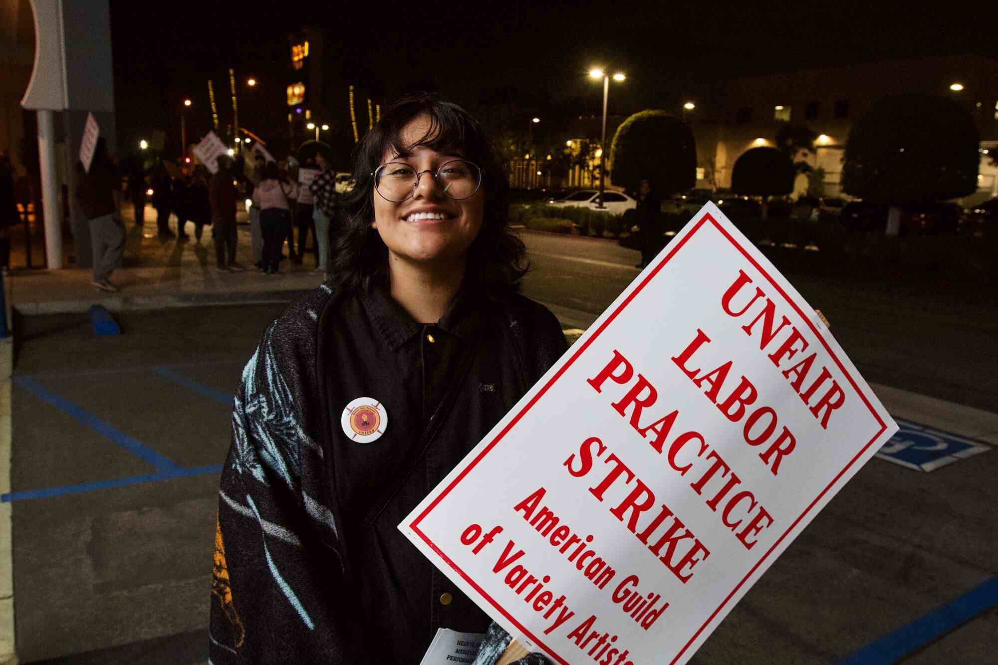 Trompeterin Lizbeth Figueroa steht lächelnd direkt vor dem Eingang und hält eine "UNFAIRE ARBEITSPRAXIS STREIK" Streikposten.