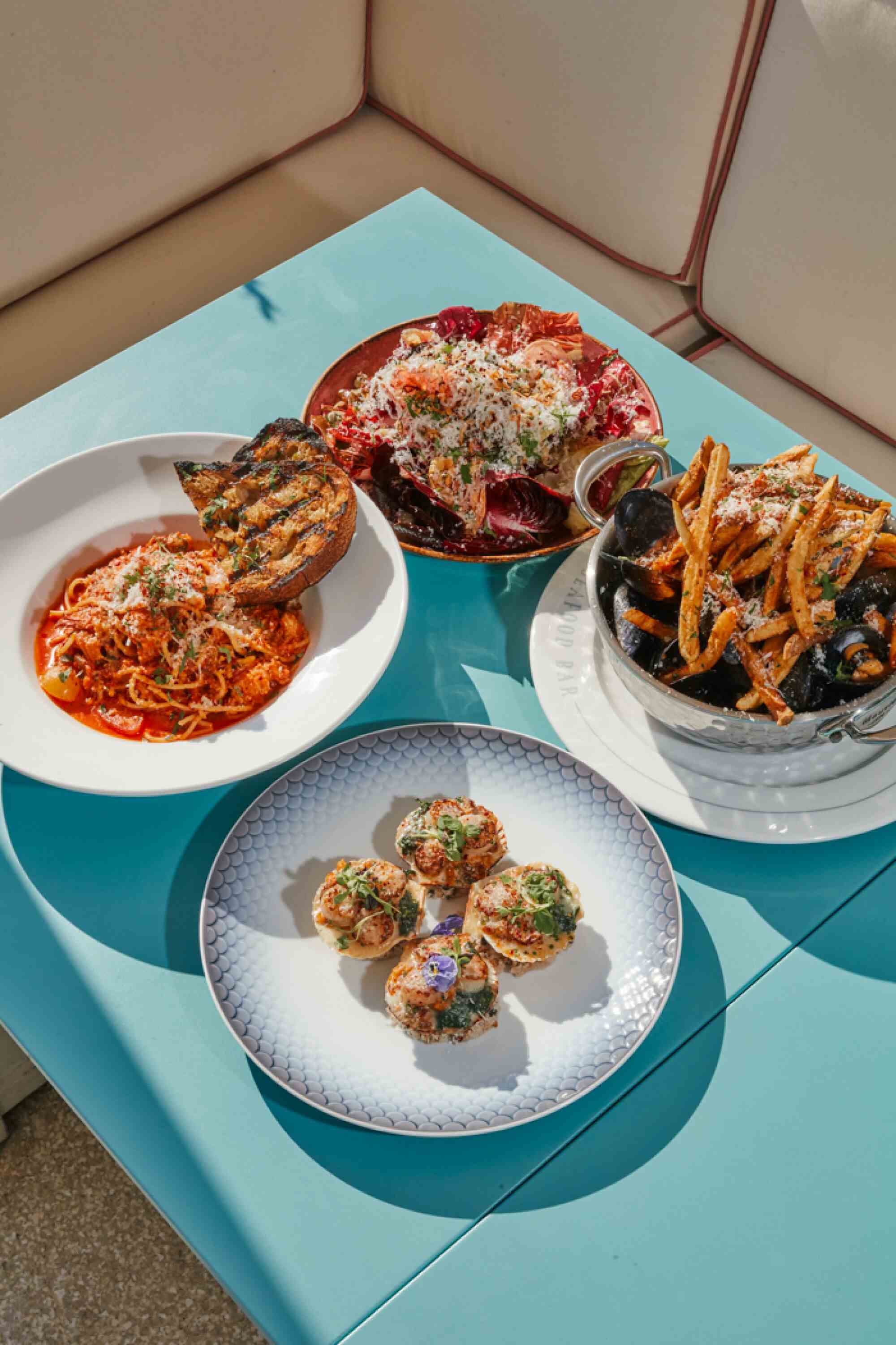 Vier Teller mit Essen auf einer türkisfarbenen Tischplatte draußen auf einer Restaurantterrasse.