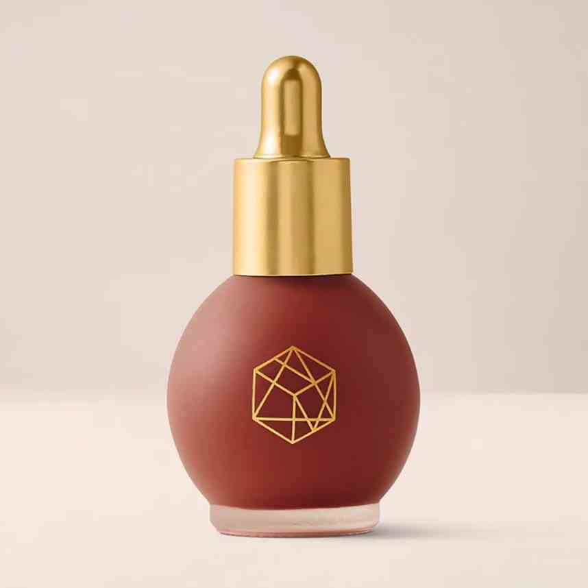 Em Cosmetics Color Drop Serum Blush rundes Fläschchen mit bräunlichem Beeren-Flüssigkeits-Rouge mit goldener Kappe auf beigem Hintergrund