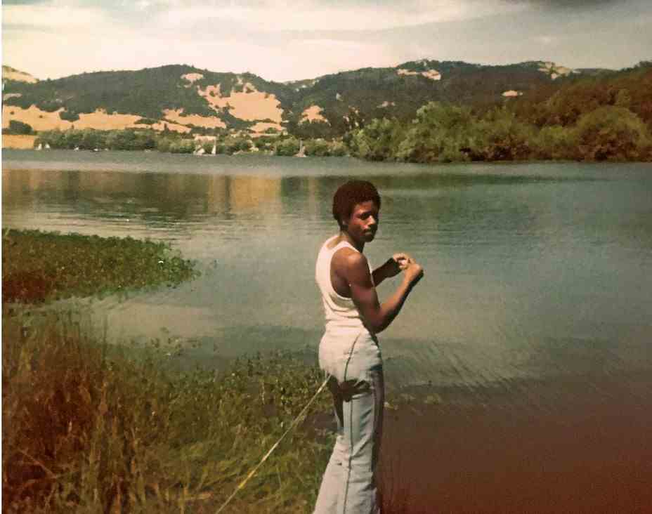 ein mann an einem see schaut in die kamera, die einen fisch hält, umgeben von bergen um die 1970er jahre.