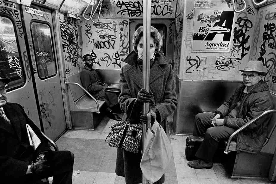 eine Frau in einem mit Graffiti besprühten U-Bahn-Wagen.  Sie starrt in die Kamera, wobei ihr Gesicht durch den U-Bahnmast getrennt ist.