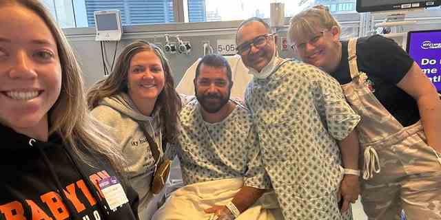 John Primavera (mit Brille) steht neben dem guten Freund und Nierenspender Tom Kenny (im Krankenhausbett sitzend) zusammen mit ihren Familienangehörigen nach ihrer erfolgreichen Transplantation an der NYU Langone Health in New York. 